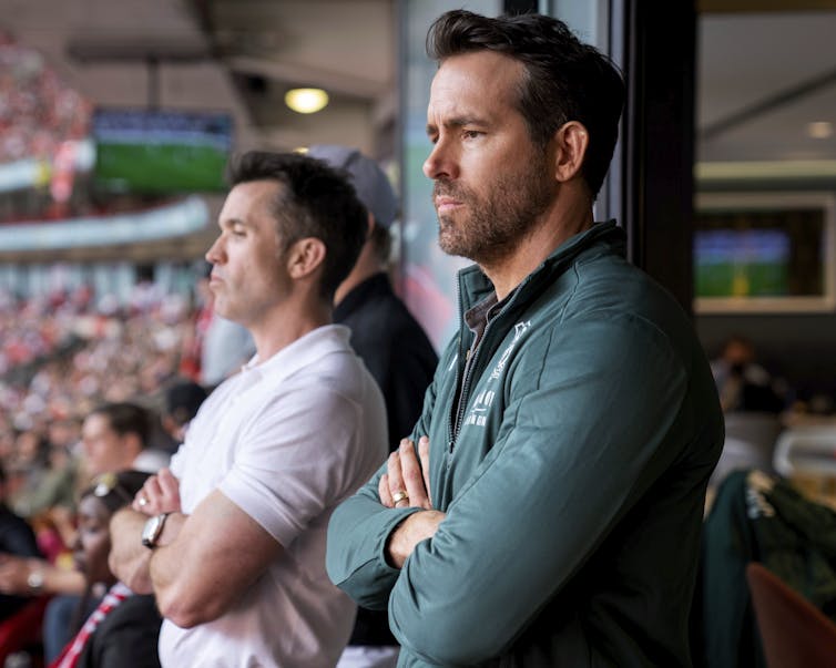 Two men with crossed arms are side by side as they watch a soccer game from the stands.