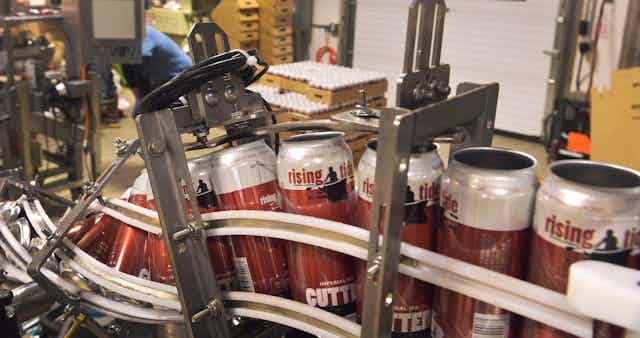 Red and white beer cans that say 'rising tide' on them, on a manufacturing conveyer. 