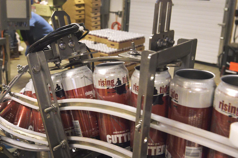 Red and white beer cans that say 'rising tide' on them, on a manufacturing conveyer.