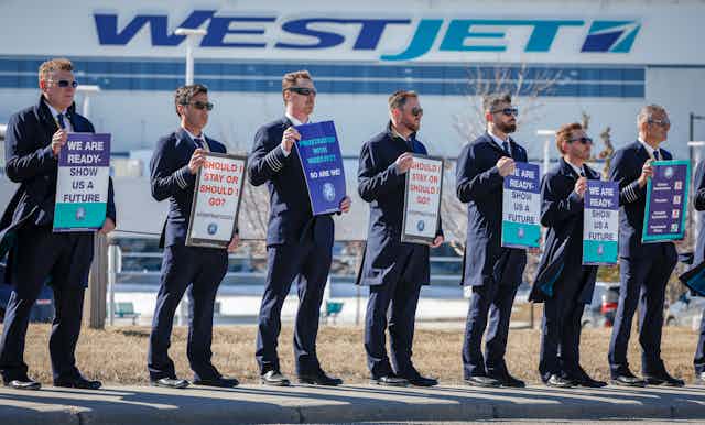 A row of men wearing navy pilot uniforms stand in front of a WestJet plan holding protest signs.