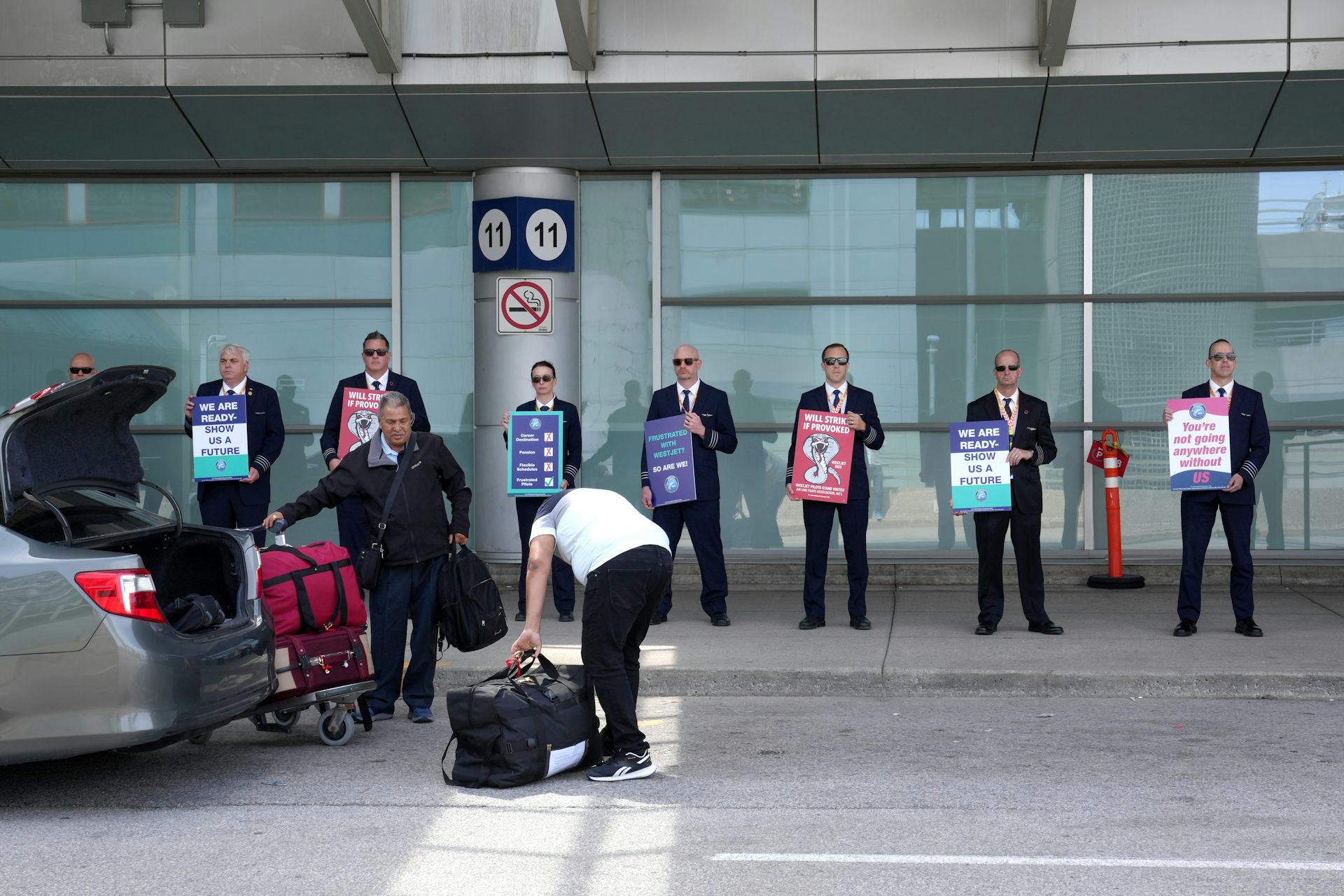A row of men wearing navy pilot uniforms hold protest signs in the background of a photo as someone unpacks luggage from the trunk of a car in front of them.