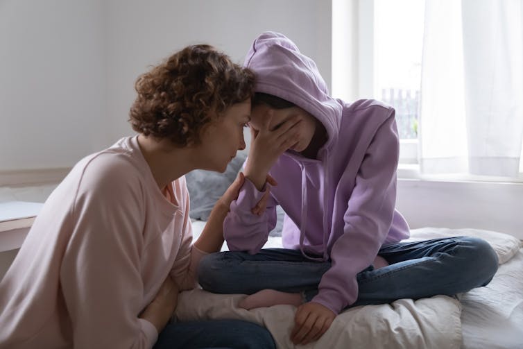 A woman comforts a teenage girl sitting on a bed.