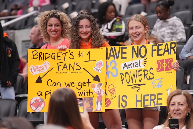 Three teenage girls hold signs supporting their team up at a basketball game.