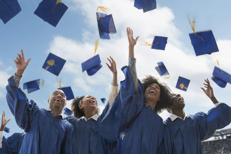 Four Black college students throw their graduation caps in the air. They are blue. So are their gowns.