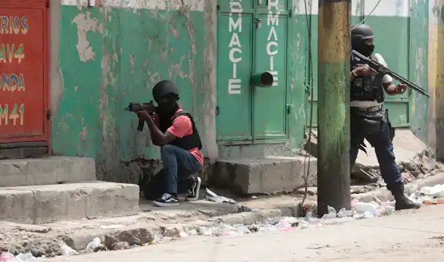 Two heavily armed police officers, one crouching and the other standing behind a pole. Both wear helmets.
