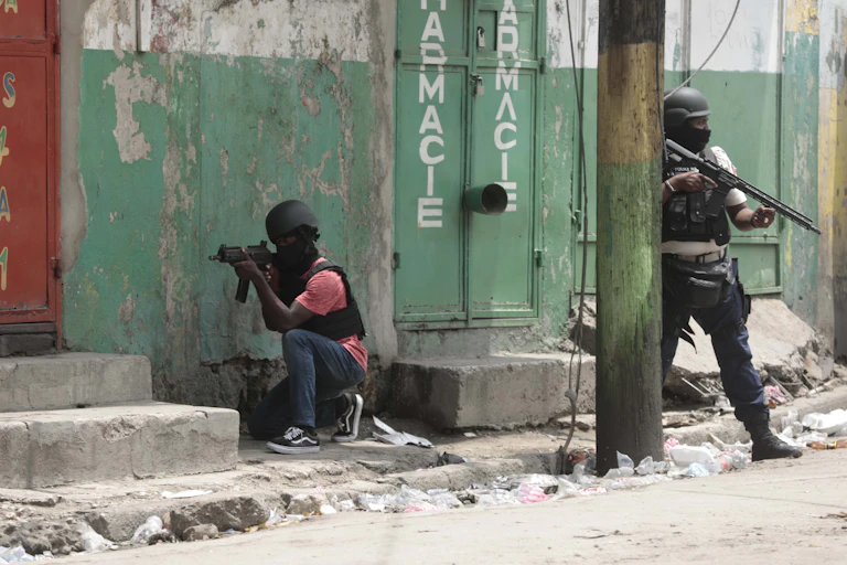 Two heavily armed police officers, one crouching and the other standing behind a pole. Both wear helmets.
