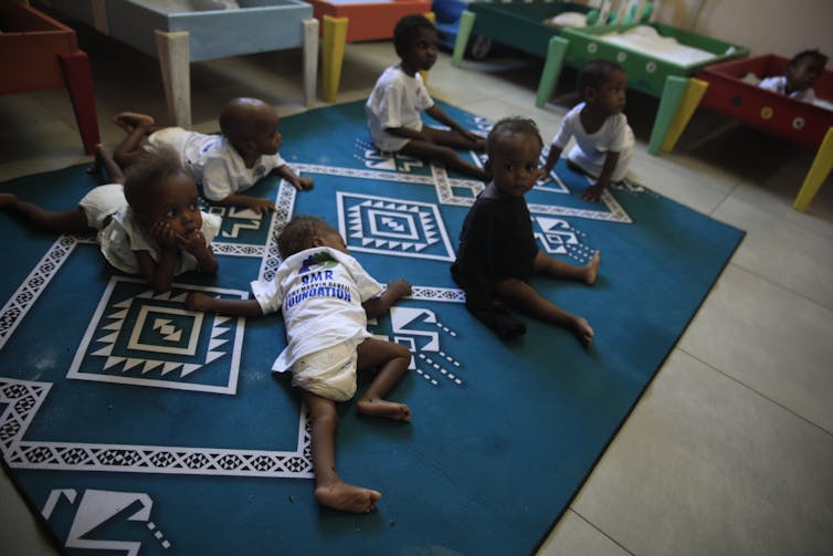 A group of toddlers play and rest on a blue and white mat.