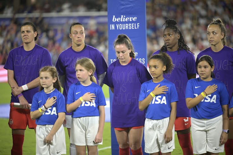 A row of women soccer players stand with their hands behind their backs. In front of them stands a row of young girls in soccer uniforms.