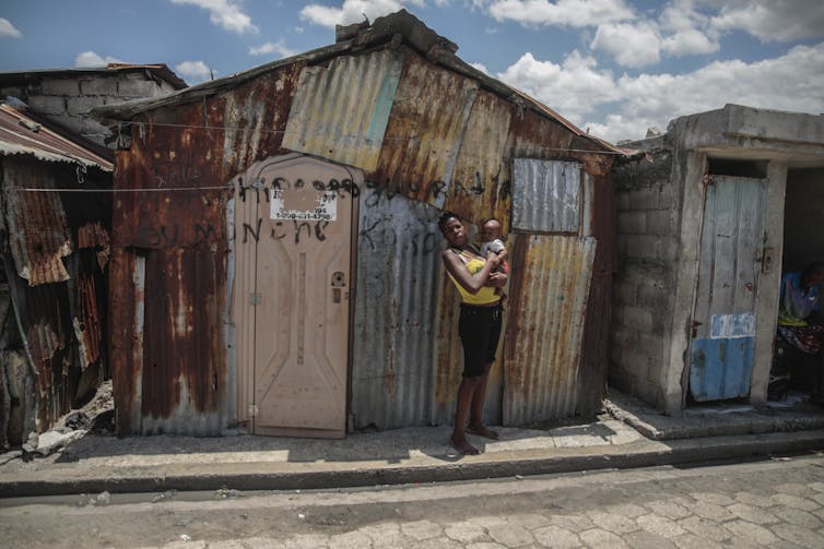 A woman holding a baby stands in front of a shack made of rusted corrogated metal under bright sunlight.
