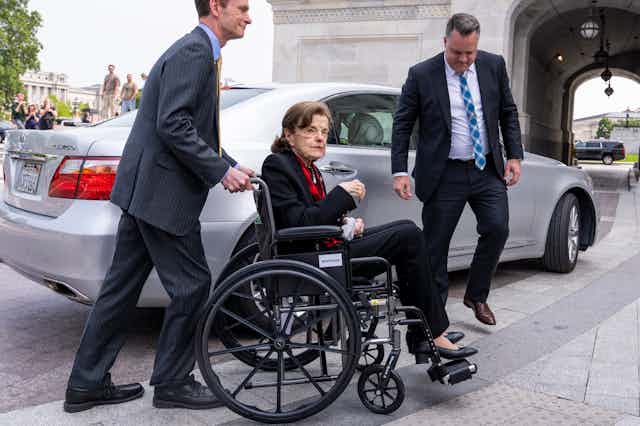 A dark-haired woman wearing a black suit, in a wheelchair near a car.
