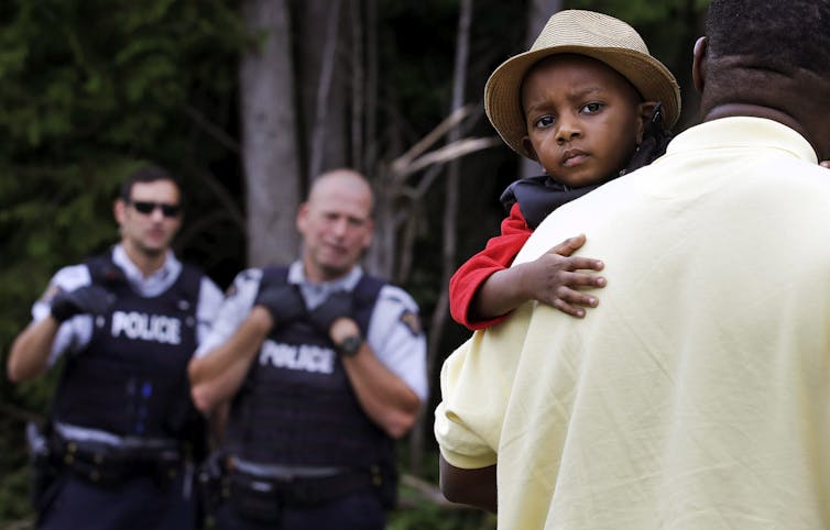 A young boy wearing a fedora is carried by his father towards two RCMP officers.