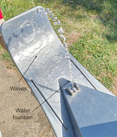 A drinking fountain with water flowing down a metal slope, exhibiting waves.