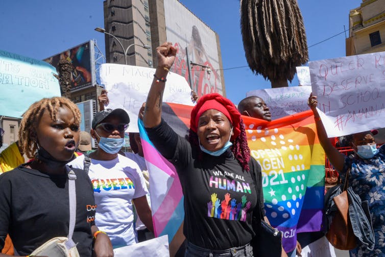 A protest with a crowd of people holding banners and rainbow flags, in front are three women, one with a raised fist.