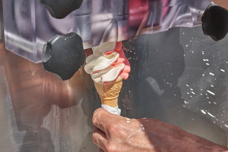 A hand holding a waffle cone under the nozzle of a machine dispensing pink and white soft serve