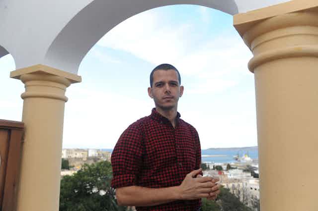 A man stands on a balcony under an arch with a view of a coastal city behind him. He holds his hands in front of him and looks directly to camera.