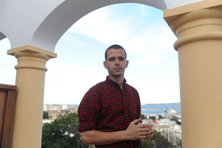A man stands on a balcony under an arch with a view of a coastal city behind him. He holds his hands in front of him and looks directly to camera.