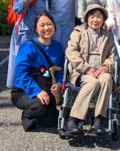 An elderly Japanese woman in a wheelchair with her granddaughter, both smiling.