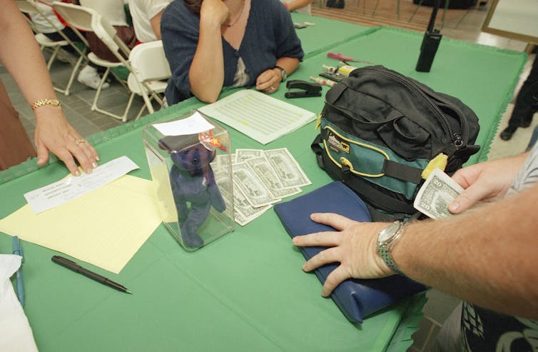 Close-up of a table with a small stuffed animal in a clear plastic case in the middle. On either side, hands exchange US dollars.