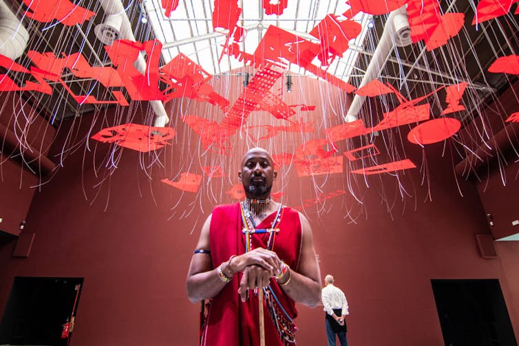 A man stands in traditional African attire in an exhibition space with red artworks suspended from the ceiling.