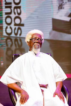 An elderly man with grey haired a white robe sits in a chair.