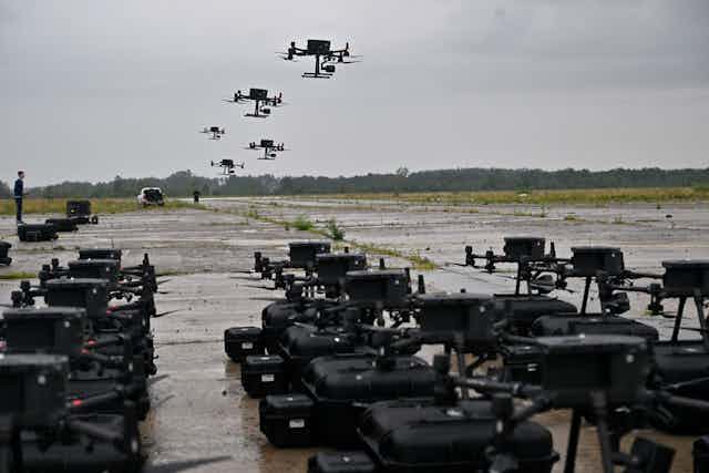 Rows of black military vehicles are seen on the ground, while about 7 drones fly above head in a grey sky.