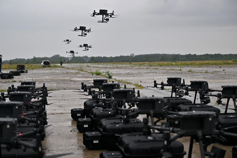 Rows of black military vehicles are seen on the ground, while about 7 drones fly above head in a grey sky.