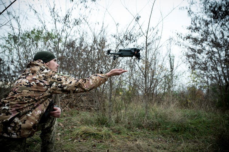 A man wearing camouflage clothing and a green hat extends his hand and a small drone flys away from him.