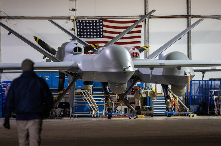 Un gran dron gris está parado frente a una gran bandera estadounidense.