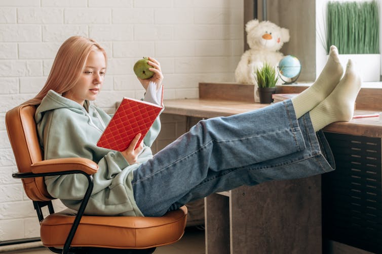 A teenage girl, relaxed, wearing jeans, and with feet up on her desk, reads a book.