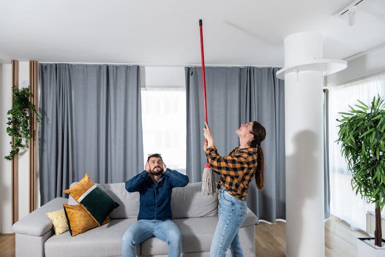 A woman banging on the ceiling of an apartment with a broom handle. A man, who is sitting on a couch in the same room, covers his ears and watches her.