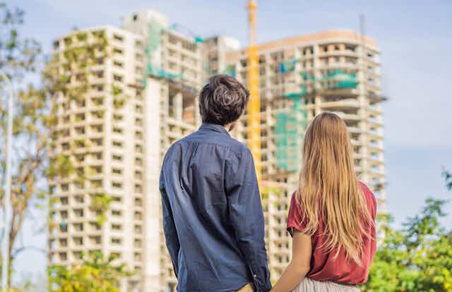 A man and a woman hold hands while looking at an apartment building under construction. They are facing away from the camera.