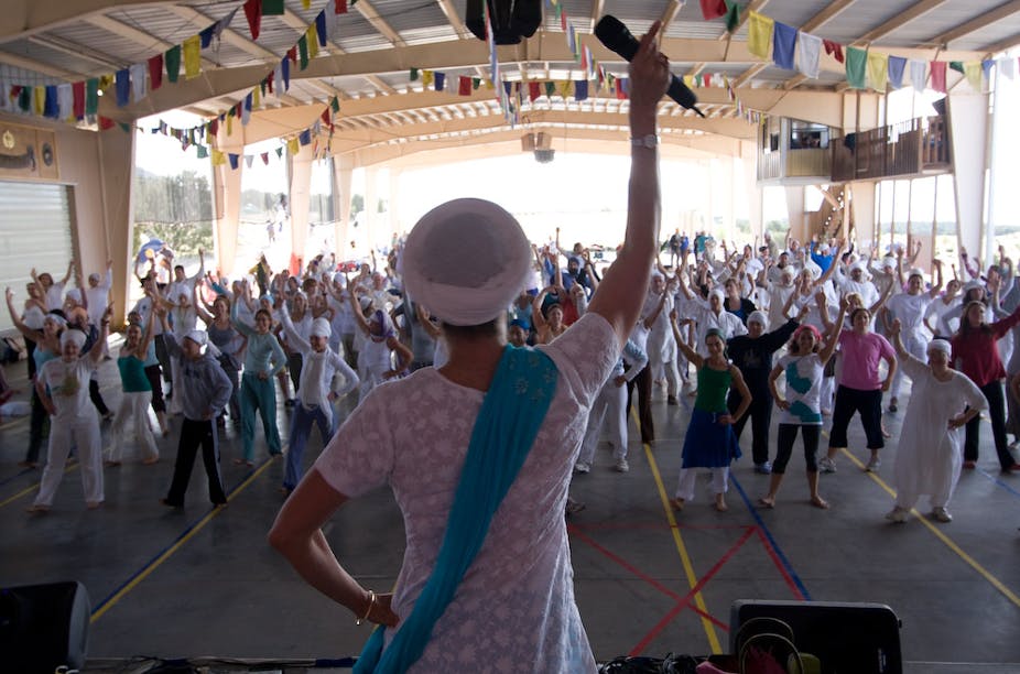 A woman wearing a white shirt raising one arm holding a microphone while leading a group exercise.