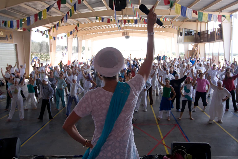 A woman wearing a white shirt raising one arm holding a microphone while leading a group exercise.