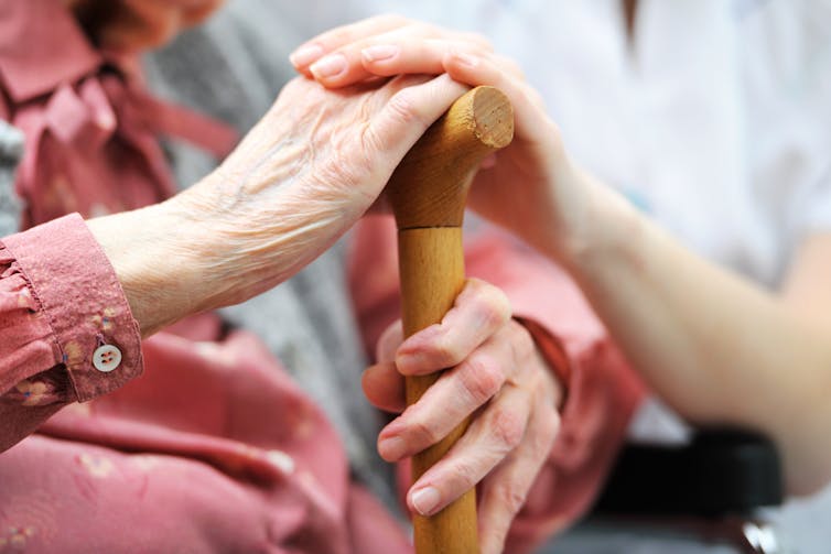 carer puts hand on older person's hand, which is hold a walking stick