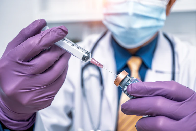 A doctor wearing purple gloves and a mask pokes a needle into a vial.