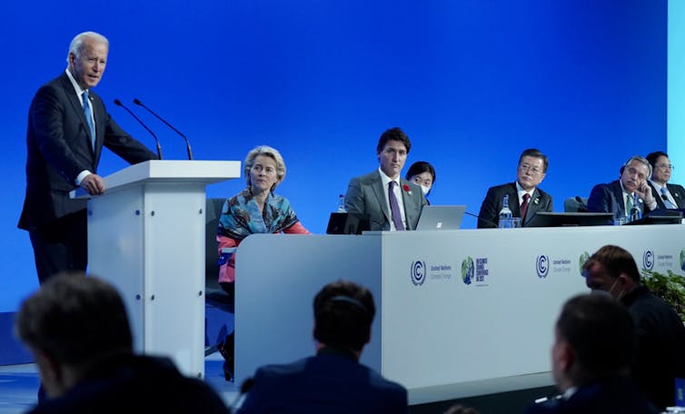 Joe Biden stands at a lectern with world leaders seated on his left.