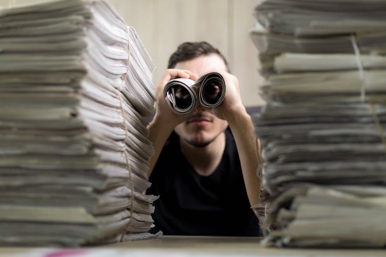 Man between piles of paper holding up two rolls to his eyes