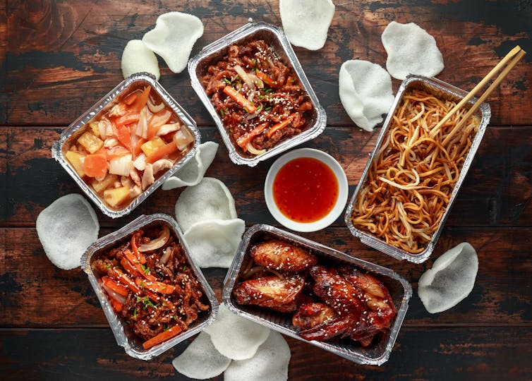 Five foil takeaway containers with colourful food inside including chicken chow mein, chilli beef and prawn crackers.