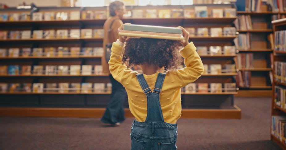 A toddler in dungarees is seen walking away from the camera, carrying books on their head in what looks like a library or book store