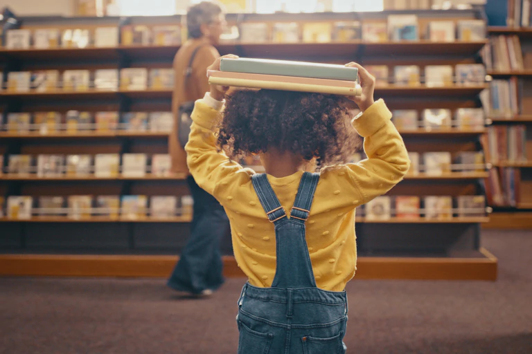 A toddler in dungarees is seen walking away from the camera, carrying books on their head in what looks like a library or book store