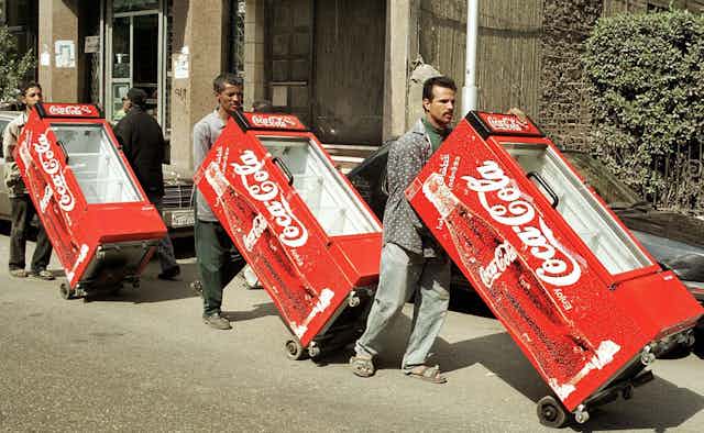Three men roll coolers with Coca-Cola logos along a city street
