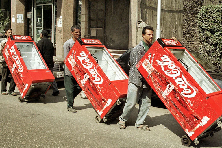 Three men roll coolers with Coca-Cola logos along a city street
