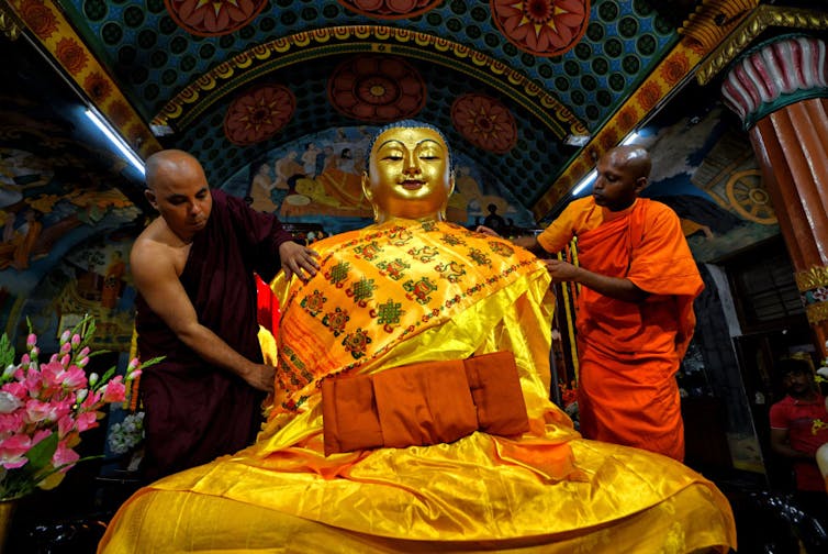 Two monks in dark red and bright orange robes carefully handle a large gold statue of a seated Buddha.