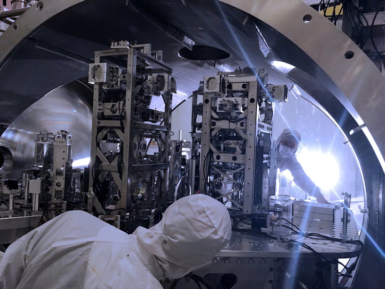 Two people in white lab outfits working on complicated machinery.