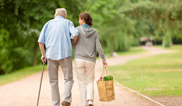 A middle-aged woman helps an older man walk while she carries a bag of groceries.