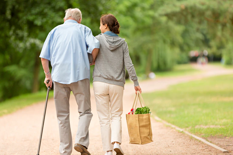 A middle-aged woman helps an older man walk while she carries a bag of groceries.
