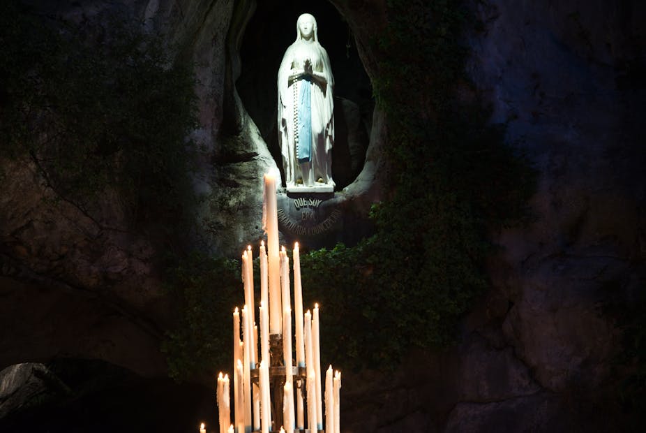 Several candles lit before Virgin Mary statue at Lourdes.