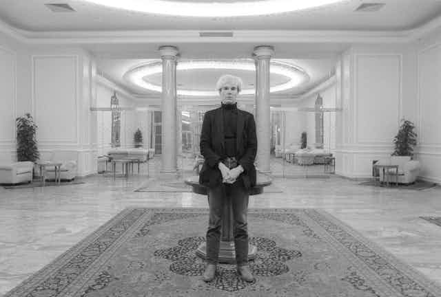 Andy Warhol, wearing a black turtleneck shirt and jacket, and dark-colored pants, stands before the camera.