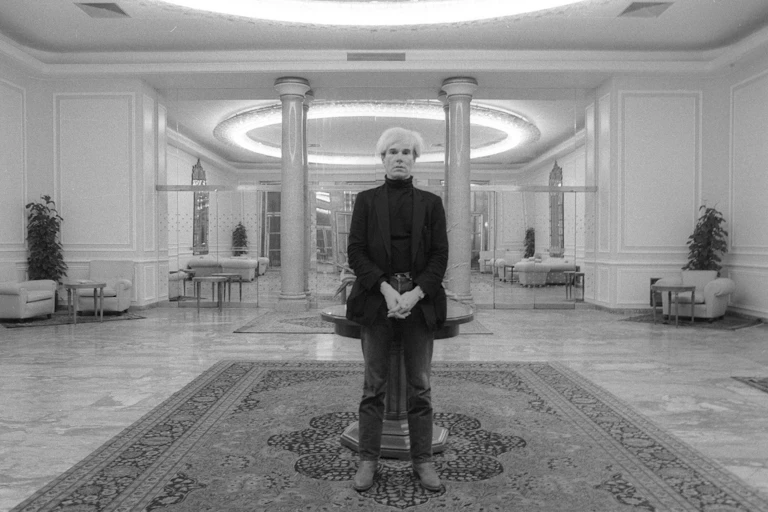 Andy Warhol, wearing a black turtleneck shirt and jacket, and dark-colored pants, stands before the camera.