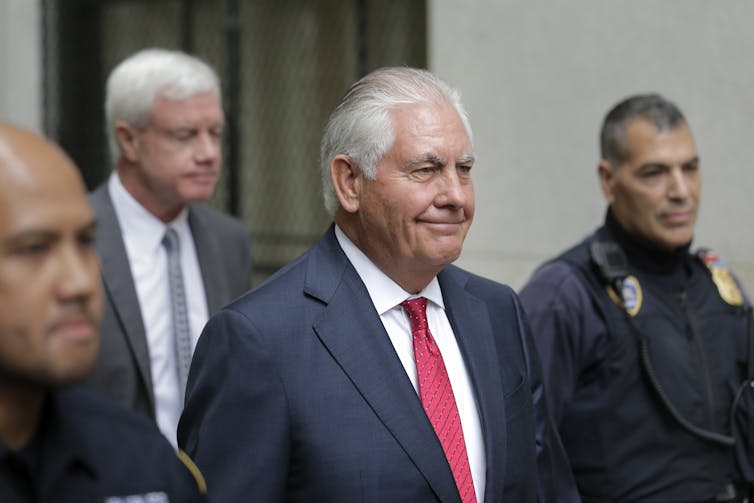 Rex Tillerson, a smiling older man in a suit and tie, walks out of a courthouse with security guards.